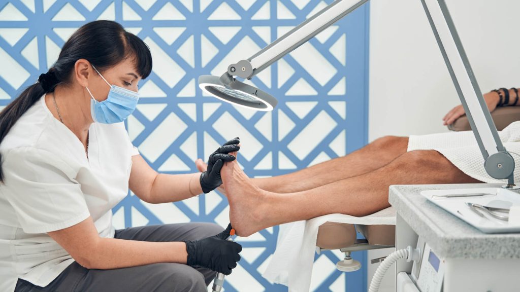 Female beautician in medical mask using electric device while removing old skin from male foot